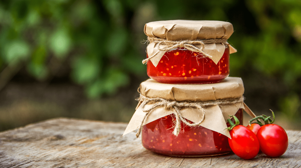 Two jars of hot sauce with seeds stacked on top of each other framed by greenery and a few tomatoes